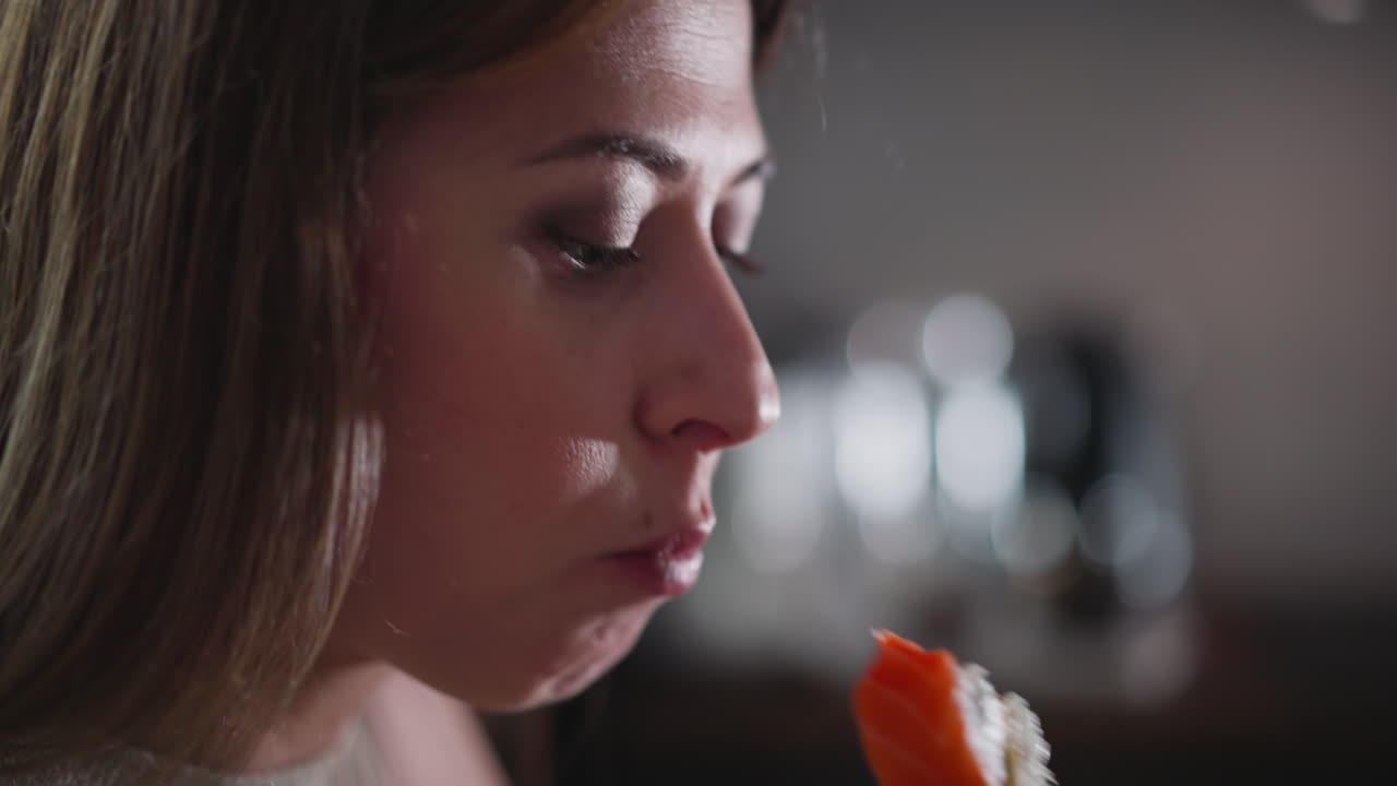Close up of business lady holding sushi with chopstick near lips while preparing to eat, natural light highlights her focused, background features soft bokeh lights from blurred modern kitchen