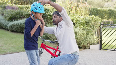 indian father helping young son adjust helmet while standing next to bicycle