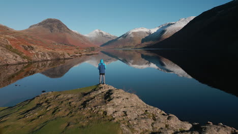 flying past hiker on lake outcrop with high water reflections and snowy mountains at wasdale lake district uk