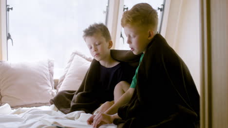 two little brothers with blanket on their shoulders sitting on bed in a campervan and reading a book