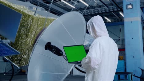 vista trasera de un científico masculino asiático que usa una computadora portátil de pantalla verde mientras está de pie con la construcción de una nave espacial