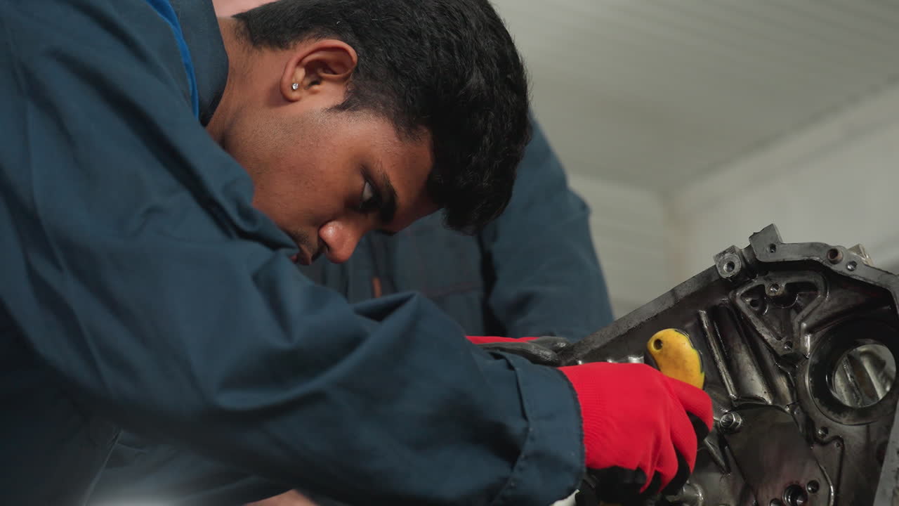 Mechanic in blue uniform wearing red gloves focusing intently on car engine, using a yellow-handled tool in an automobile workshop, demonstrating precision while working on mechanical components