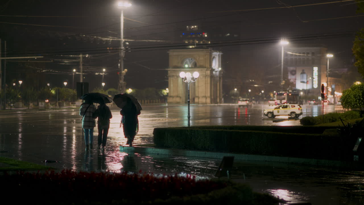People navigate near city center at night under umbrellas as rain pours down. Triumphal Arch on background. Chisinau, Moldova