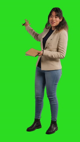 vertical video full length studio shot of female teacher with notebook talking to class standing against green screen 1