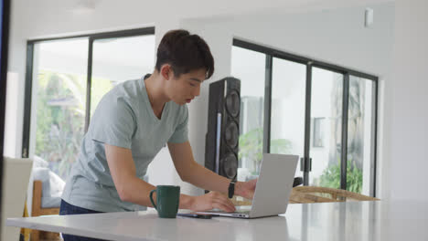 Asian-male-teenager-using-laptop-and-wearing-headphones-in-living-room