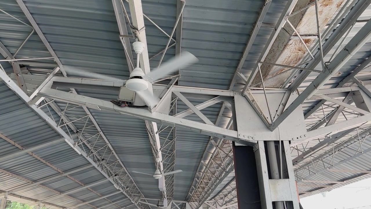 Wide shot of multiple ceiling fans operating inside an Indian railway station with large metal roof structure industrial beams and public transport infrastructure.