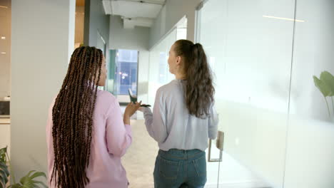 walking in office hallway, two women discussing work and holding smartphone