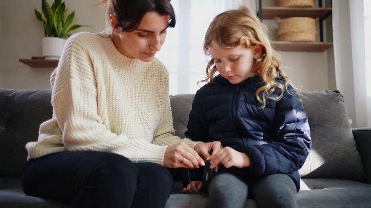 Leaning, woman guiding girl's hands adjusting black case on sofa to secure connection, navy puffer