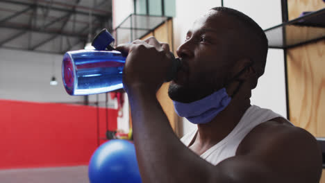 african american man wearing lowered face mask drinking at gym