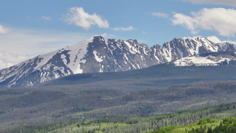 Very-closeup-aerial-view-of-beautiful-Colorado-mountain-range-with-snow-capped-peaks-on-a-sunny,-blue-sky-day-in-the-summer-with-green-fields,-trees,-and-mountain-homes