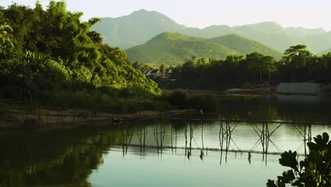 Three People Walking Across a Wooden Foot Bridge Above the Mekong River. The Scenery Features Pictureseque Mountains and Hills with the River Very Calm.