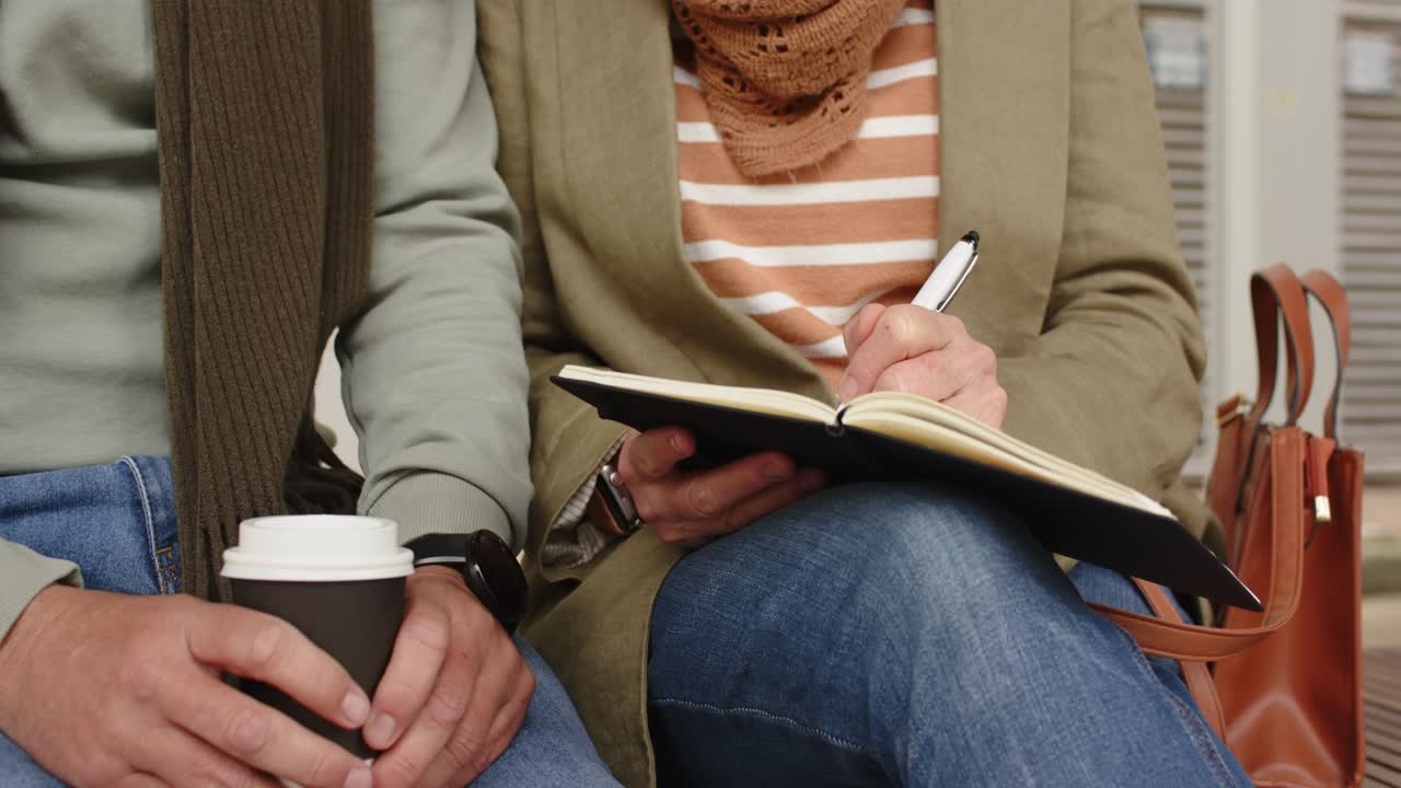 Coppia di anziani seduta su una panchina all'aperto, la donna scrive su un quaderno con una penna, l'uomo tiene in mano una tazza di caffè
