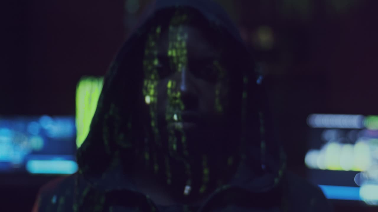 Portrait Of The Young Male Hacker In A Hood Looking Straight To The Camera With Projected Code Numbers And Characters On His Face In The Dark Room Full Of Computers