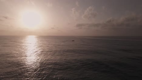 Luftaufnahme,-Die-In-Richtung-Eines-Silhouettierten-Bootes-In-Der-Ferne-Fliegt,-Das-Bei-Sonnenuntergang-Auf-Ruhigen-Meereswellen-Reist