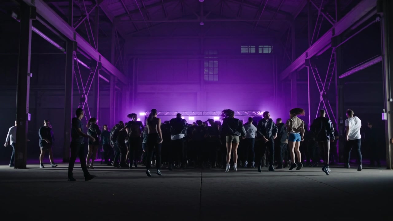 Responding to purple backlighting, crowd moving toward lit stage at hangar under truss in shorts