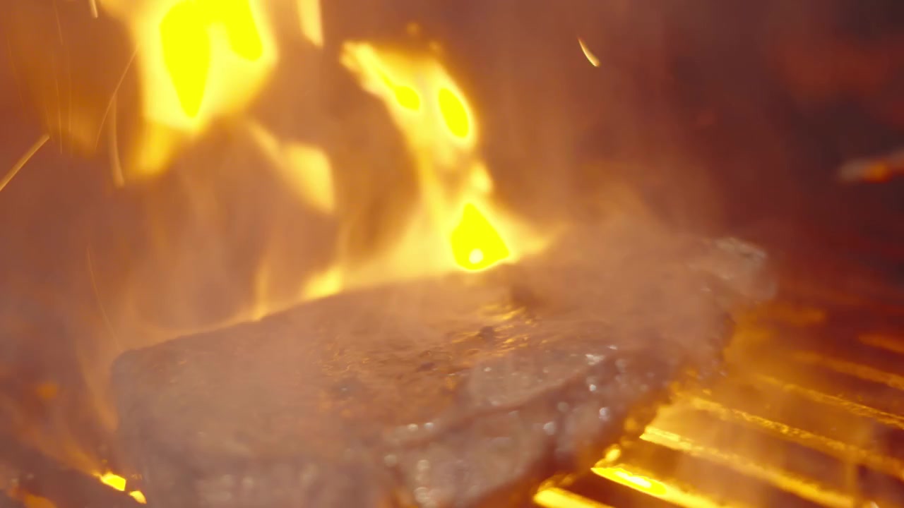 Chef che gira una bistecca di controfiletto sul barbecue con fiamme che erompono dal basso mentre la carne sfrigola sulla griglia calda.