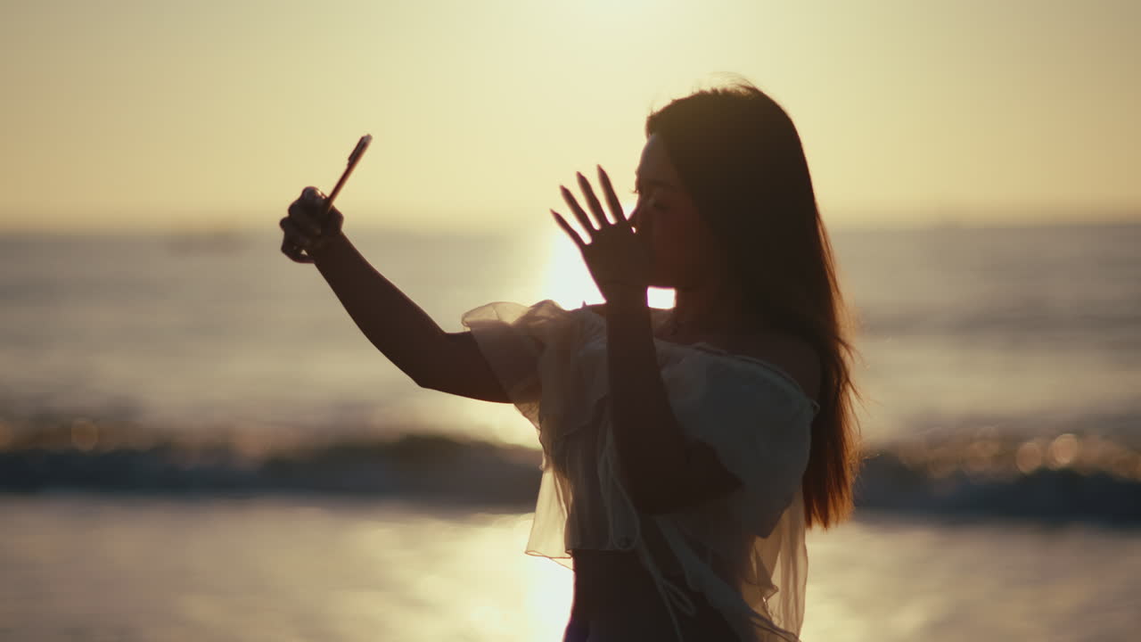 Ung kvinna tar en selfie med smartphone på stranden under en glödande solnedgång, i asiatisk influencer-stil