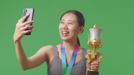 close up of asian woman with a gold medal showing a gold trophy to the video call on smartphone on green screen background in the studio