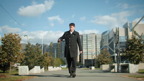 businessman in black coat walking confidently outdoors in urban setting, retrieving phone from pocket to answer incoming call, background includes greenery, modern architecture, and vibrant city