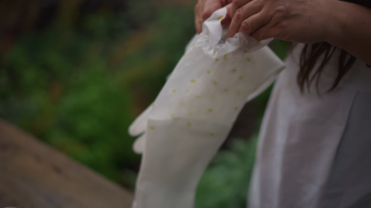White woman placing lace gloves outdoors on weathered wooden table, brass basket with petals nearby, delicate embroidered cuffs, soft daylight and lush greenery, bridelike hostess arranging heirloom