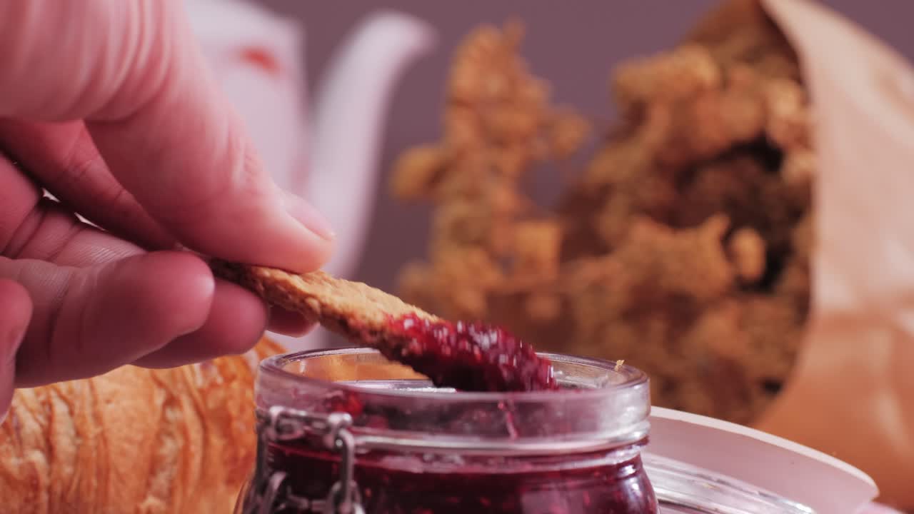 Dipping a cookie into a jar of jam with a croissant in the background