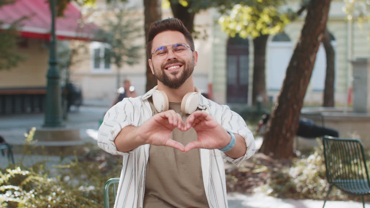 Smiling caucasian young man makes symbol of love showing heart sign to camera on city town street