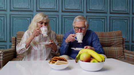 Senior-couple-holding-drinking-tea-talking,-laughing,-smiling,-kissing,-sitting-on-couch-at-home