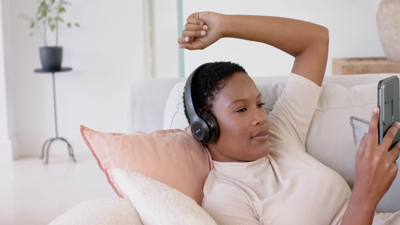 Donna afroamericana felice sdraiata sul divano, con le cuffie e che usa lo smartphone, al rallentatore