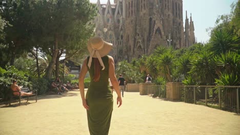 stunning video of a young caucasian female in a green dress with a knitted hat, walking to incredible landmark building - sagrada familia, barcelona