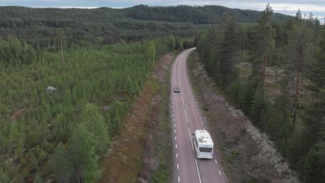 aerial tracking shot behind camper van crossing along forest road, surrounded by high pine trees, in mountainous environment