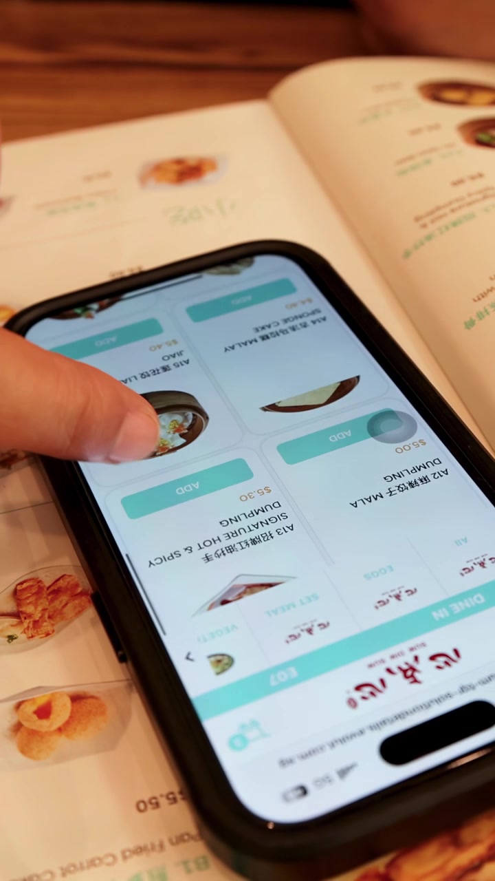 Hand interacts with smartphone food ordering app, selecting dishes in a well-lit café environment