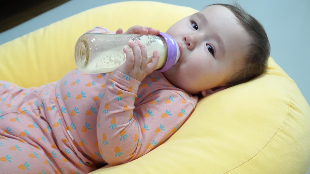 Bambina caucasica che tiene il biberon e beve latte artificiale mentre è sdraiata su un cuscino giallo - primissimo piano