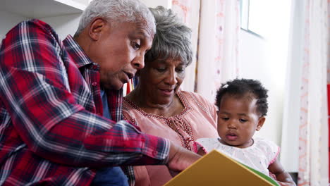 grandparents sitting on sofa reading book with baby granddaughter at home