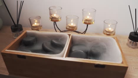 close-up of black massage stones in a wooden box emitting steam with candles in the background