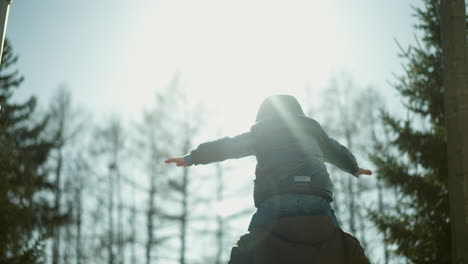 a father carries his son on his shoulders as they walk down a tree-lined path, the child stretches his arms wide, appearing cautious, while sunlight reflects off them