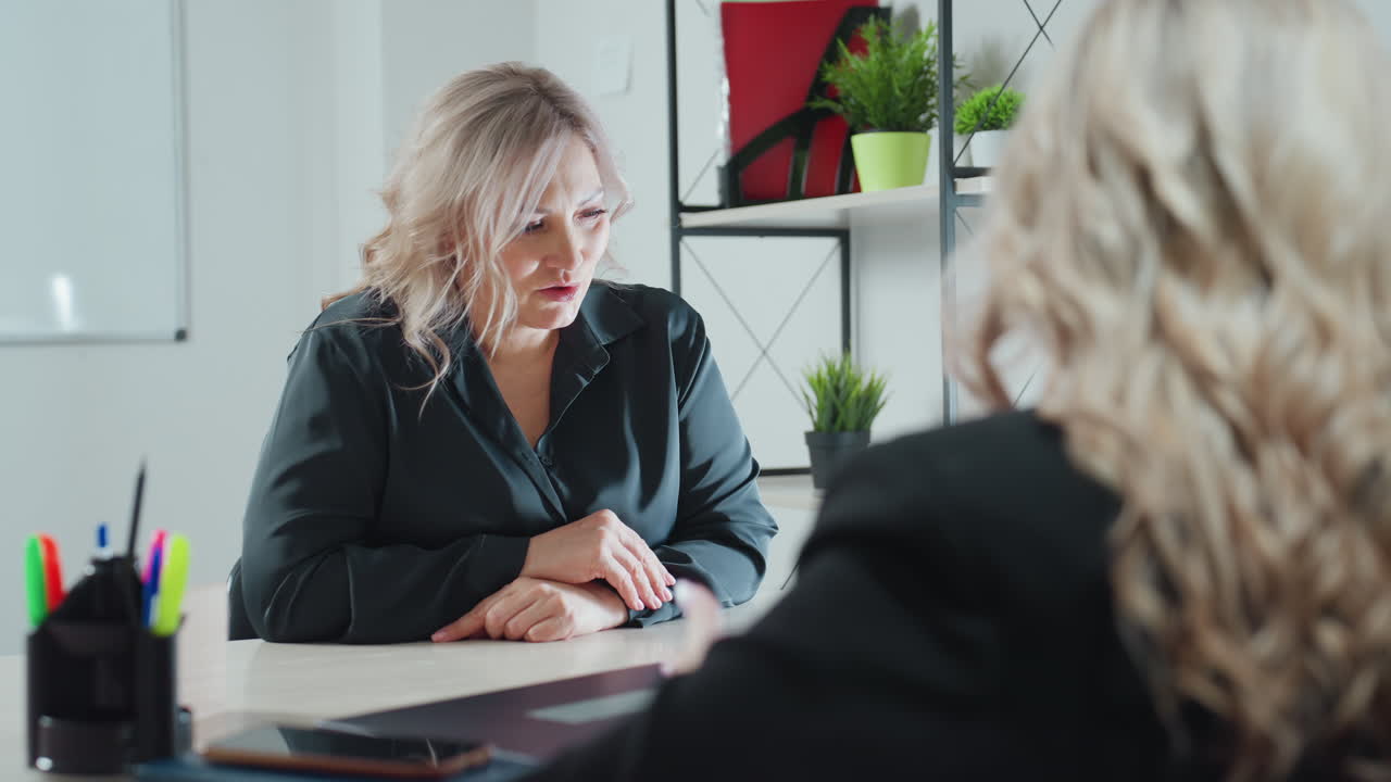 ongoing business meeting between two women seated across office desk engaged in professional conversation with focused expressions tablet notebook and pen holder visible