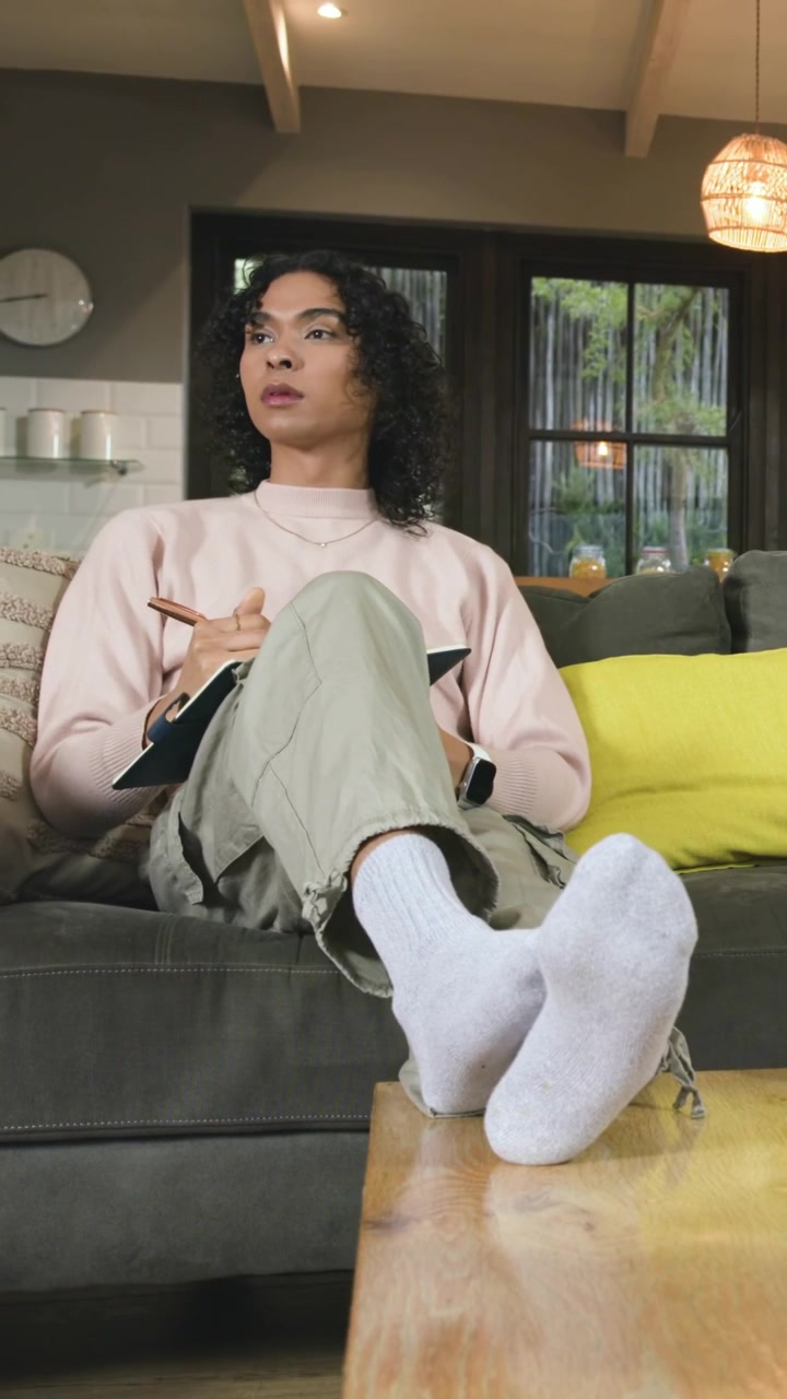 Video verticale: Adulto/a in felpa rosa appoggiato/a all'indietro con un ginocchio alzato, che scrive un diario sul divano con un quaderno.