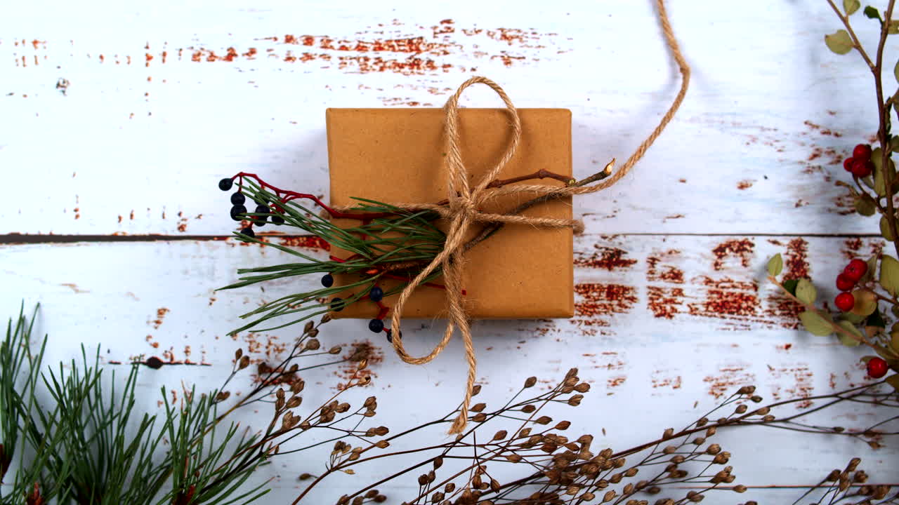 Close-up of female hands with polished nails, presenting a beautifully wrapped gift adorned with natural pine and berries. Perfect for festive promotions, DIY, and seasonal holiday themes. Flat lay.