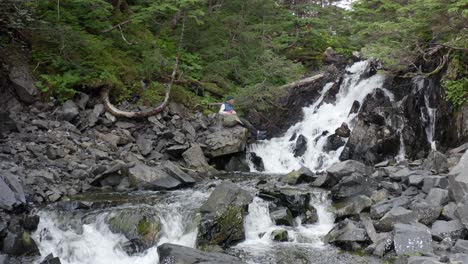 el viajero mochilero se sienta frente al agua en cascada entre rocas cubiertas de musgo en el parque forestal en alaska