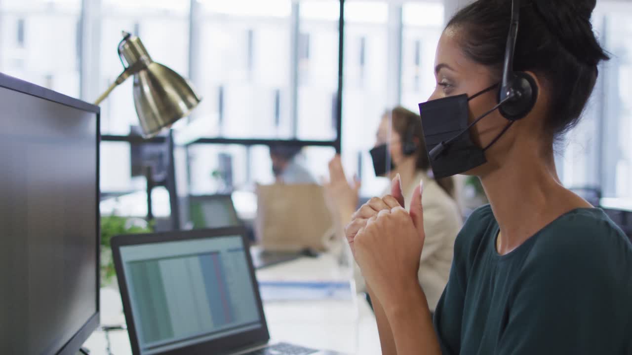 컴퓨터 를 사용 하고 전화 헤드 ⁇  을 착용 하여 이야기 하는, 책상 에서 얼굴 마스크 를 쓰고 있는 혼합 인종 사업가