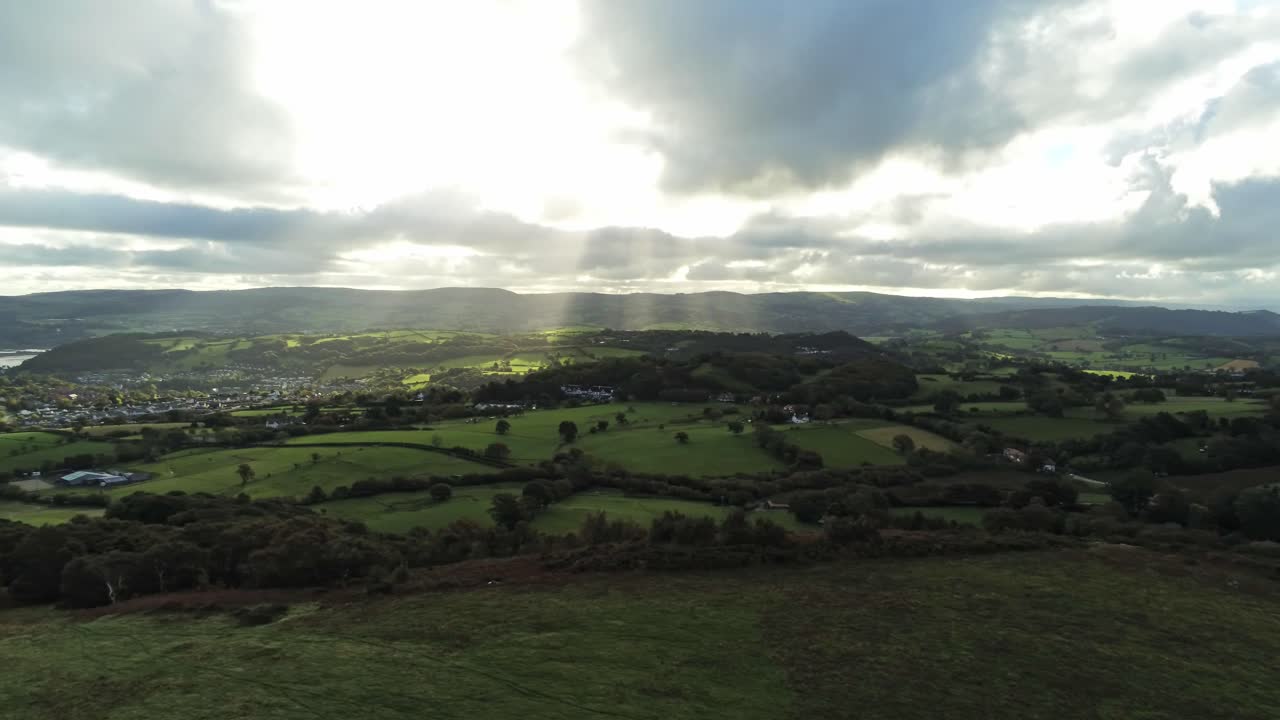 Raggi di sole che si muovono attraverso un cielo coperto sopra un paesaggio rurale agricolo britannico al mattino, con lenta carrellata aerea a sinistra