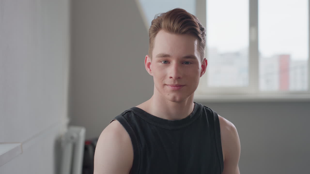 Portrait of young man in black sleeveless top sitting indoors near window, gazing forward with confident expression, natural light enters through slightly blurred window behind