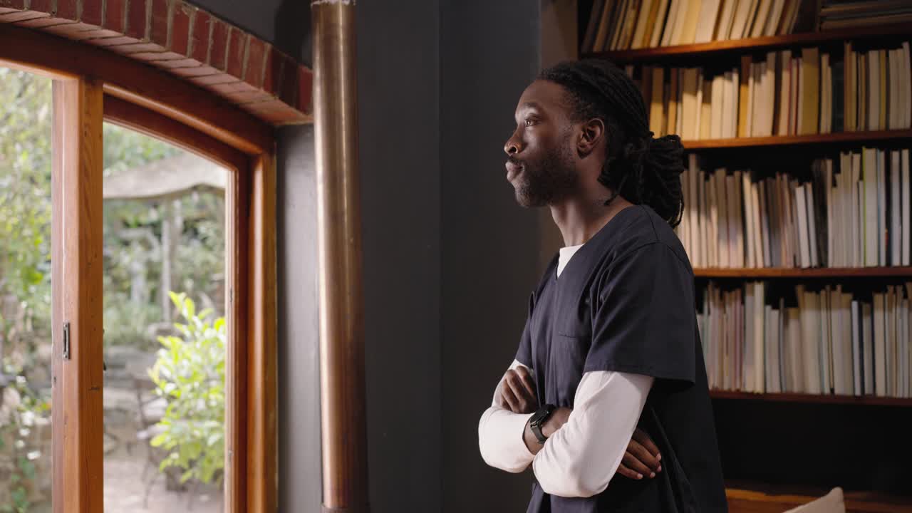 Uomo afroamericano adulto che entra da sinistra in divisa medica blu scuro, fermandosi alla finestra nella biblioteca di casa e sorridendo.