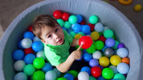 cute toddler boy sitting in the pool with ball. kid holds a red ball and pokes it with finger. top view.
