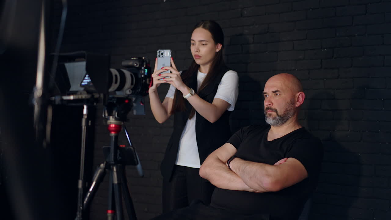 Confident serious cameraman and long-haired female assistant working in studio backstage. Focused photographer looks at camera display and girl is taking video on her phone.