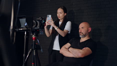 confident serious cameraman and long-haired female assistant working in studio backstage. focused photographer looks at camera display and girl is taking video on her phone.