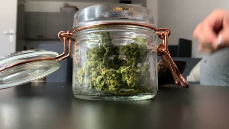 close up of a jar full of weedon a table, and a girl rolling a joint in the background