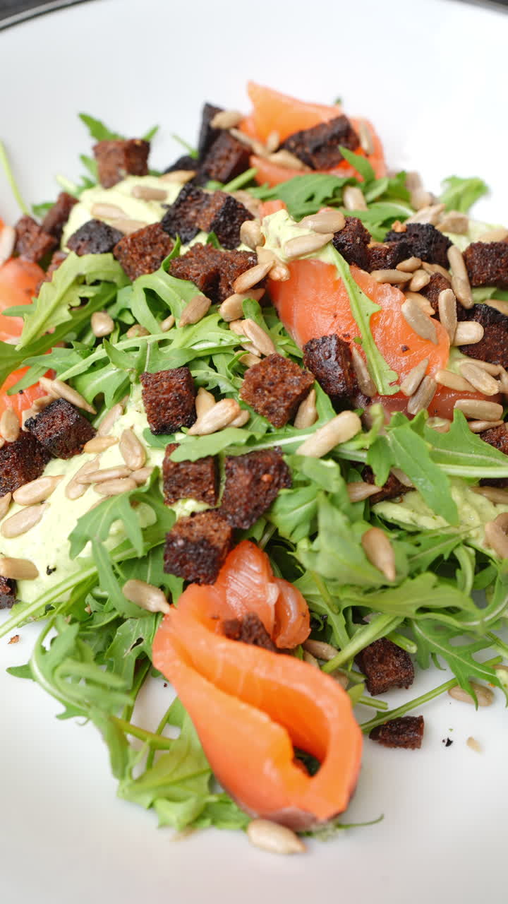 Vertical footage Gourmet salmon salad featuring fresh arugula, crispy rye croutons, sunflower seeds, and creamy sauce elegantly plated on pristine white surface