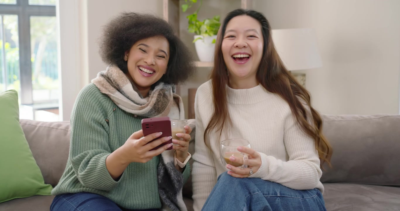 Diverse female friends sitting on couch at home showing maroon phone sharing joke with clear mugs