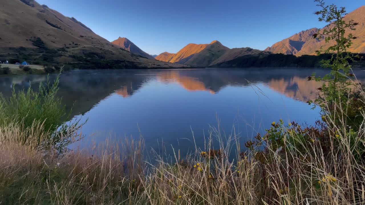 Bjerge og blå himmel spejler sig i det rolige vand ved Moke Lake med tidlig morgentåge